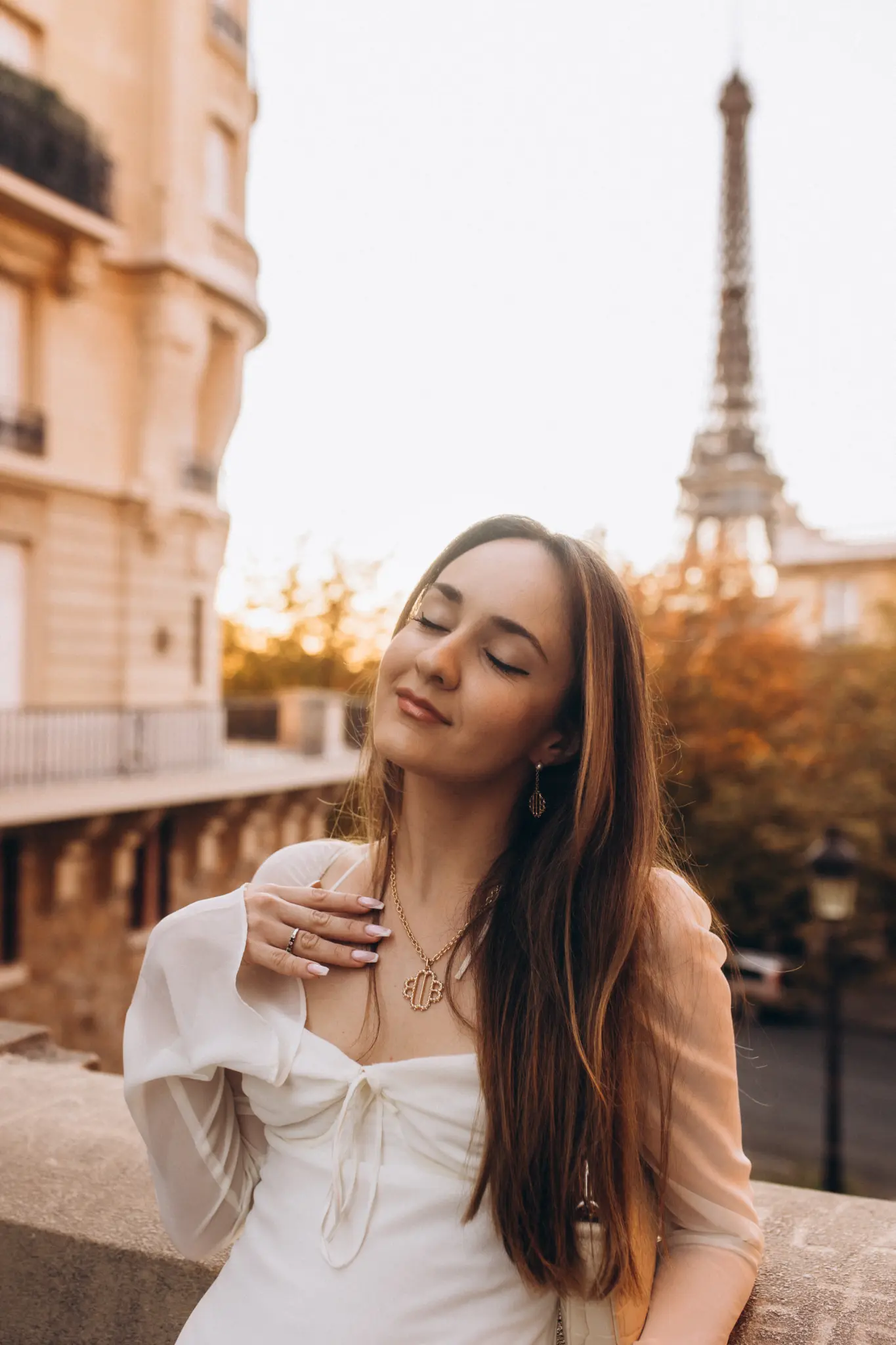 A woman in a white dress at golden hour, eyes closed, by a stone balustrade with the Eiffel Tower blurred behind her.