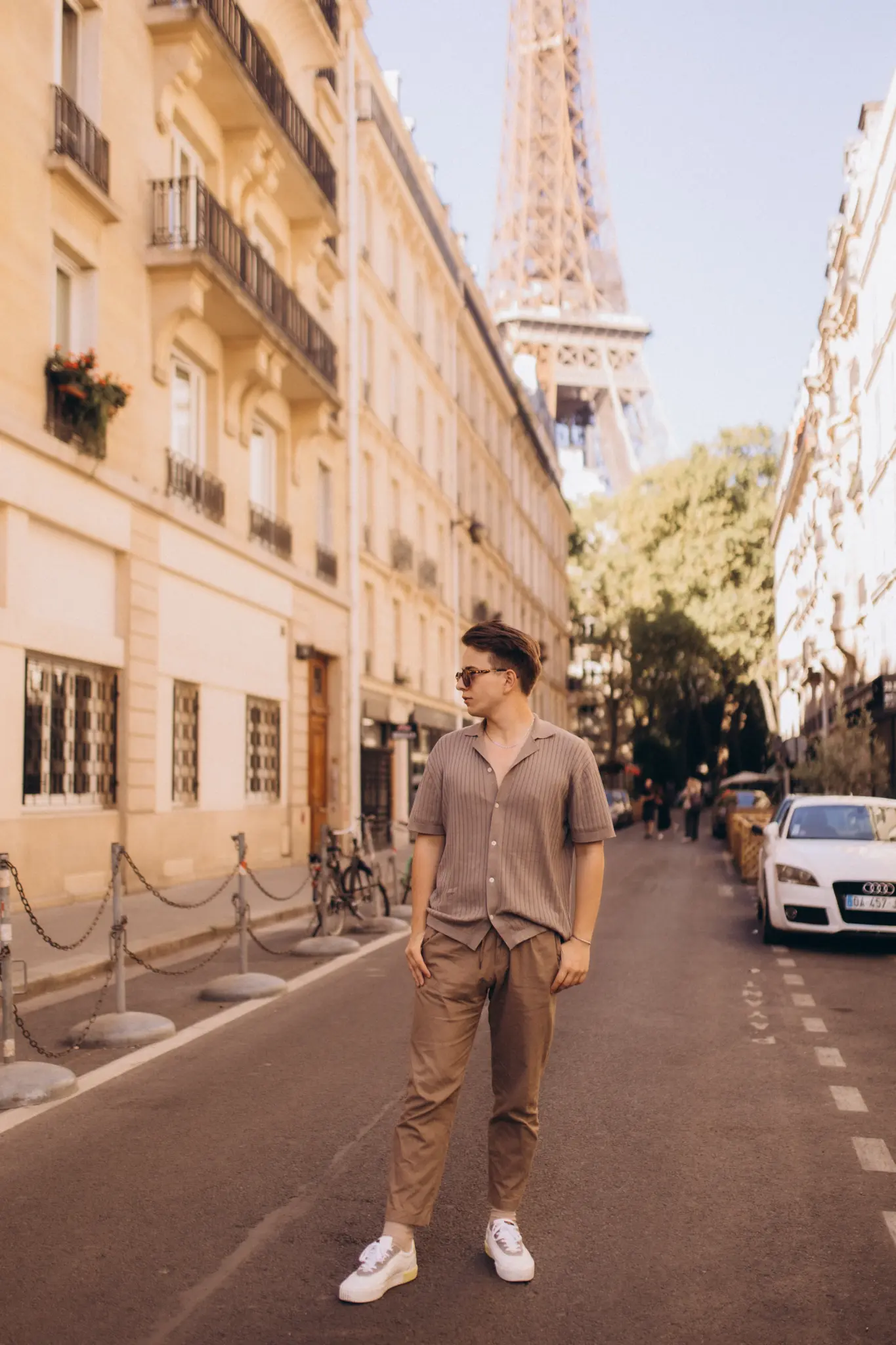 A young man in a beige knit shirt standing on a quiet Parisian street, the Eiffel Tower rising between the buildings behind him.