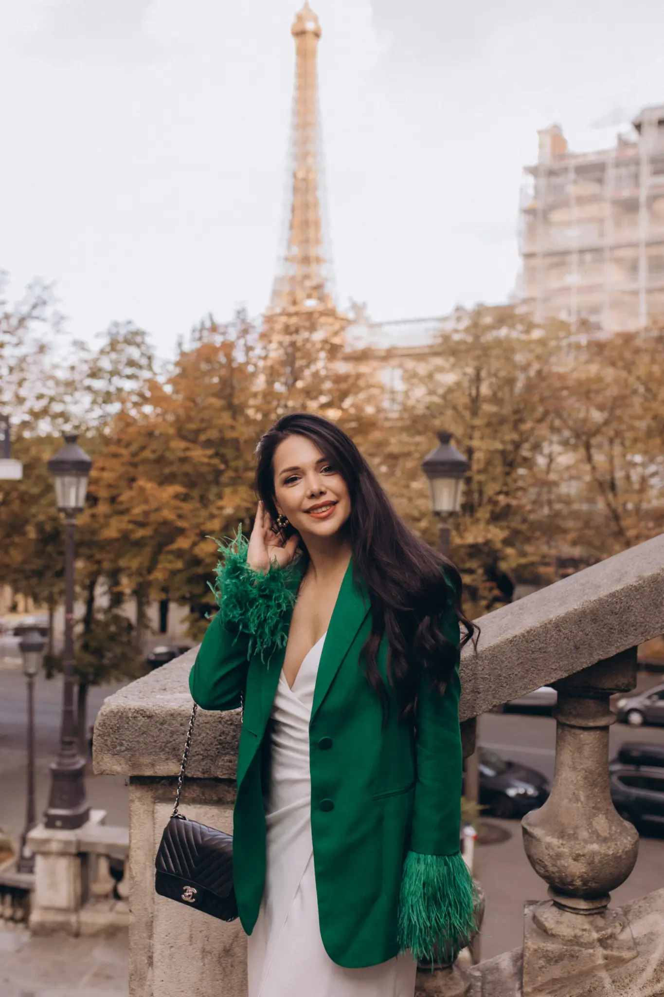 Paris portrait photoshoot, a woman in a green feather-trimmed blazer and white dress smiling by a stone balustrade with autumn trees and a famous iron landmark rising behind her