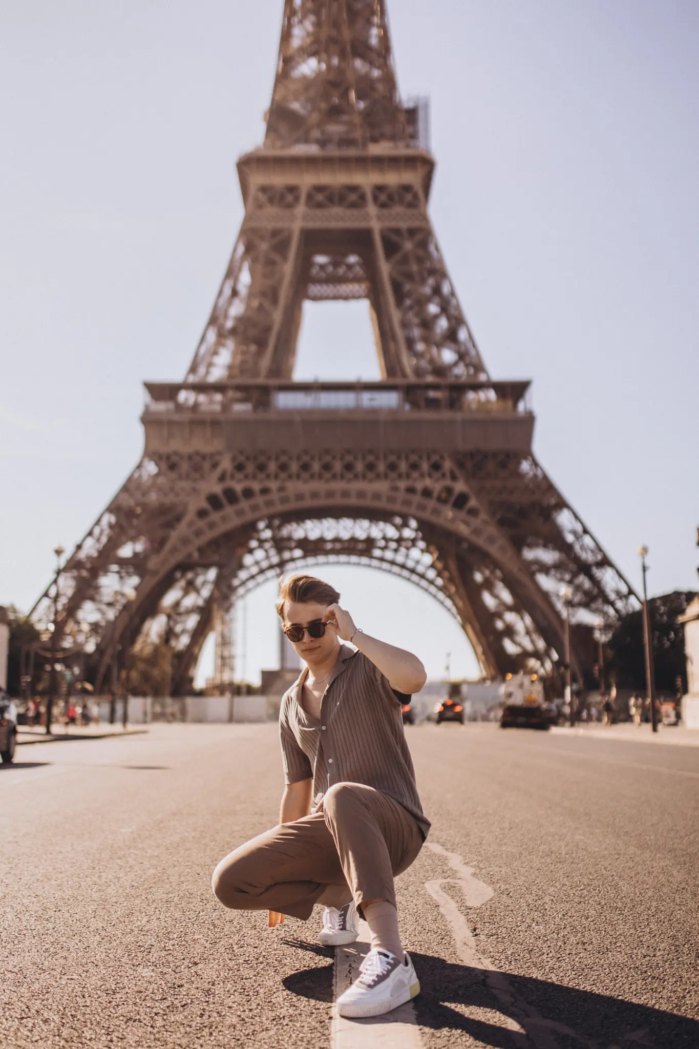 A young man crouching on an open esplanade directly beneath the Eiffel Tower, adjusting his sunglasses in bright daylight.