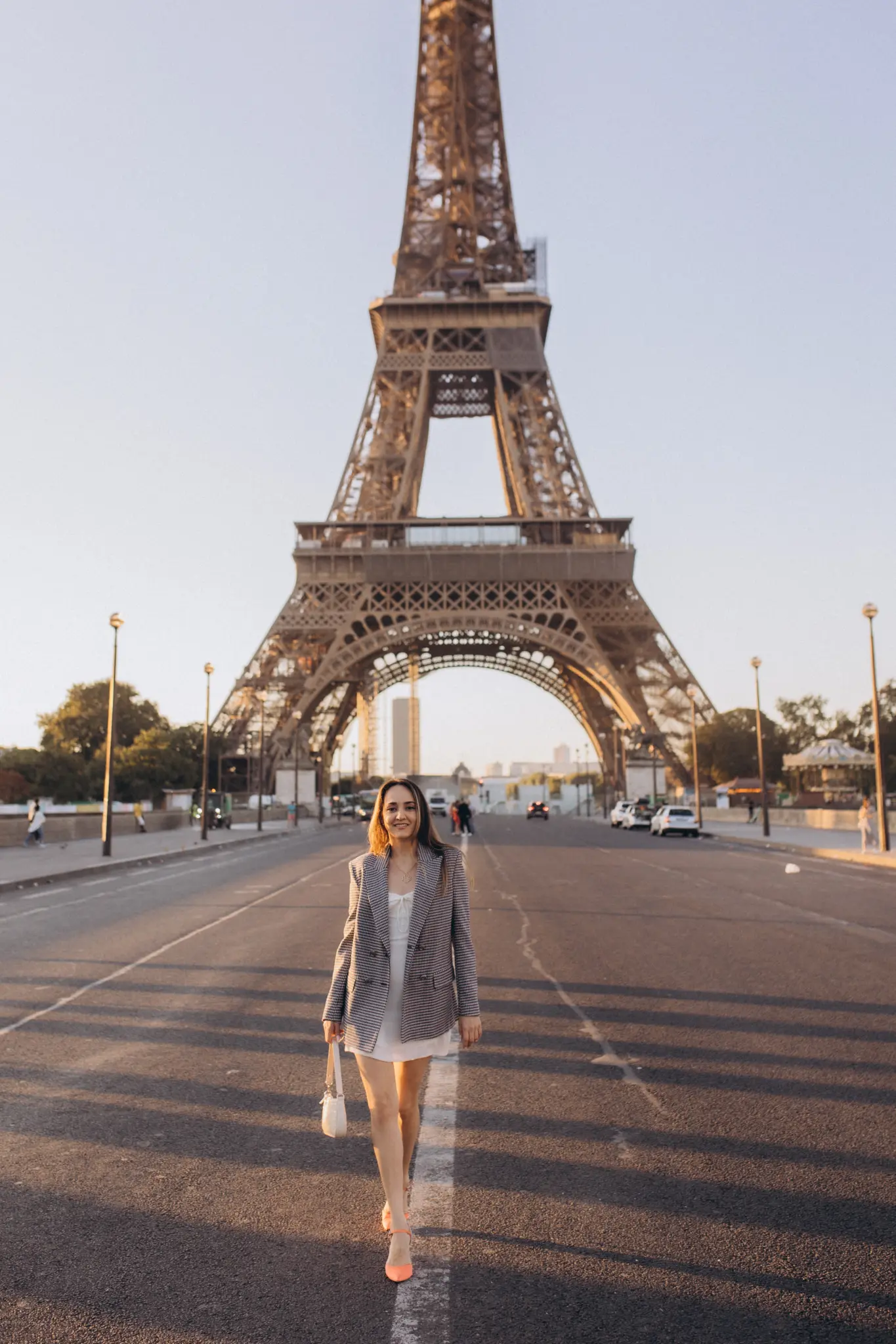 A woman in a white dress and checked blazer walking down an empty avenue straight toward the Eiffel Tower.