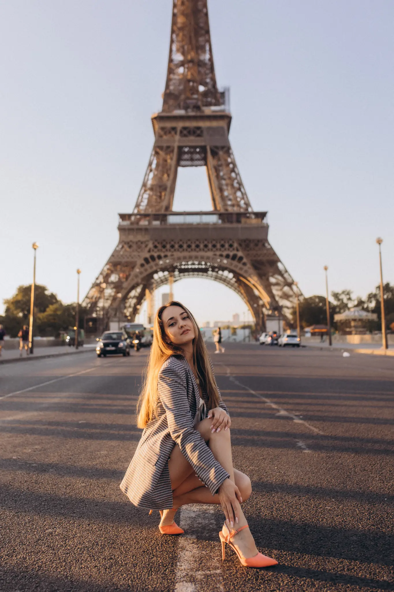 A woman in a checked blazer crouching on an empty avenue at golden hour, the Eiffel Tower filling the frame behind her.