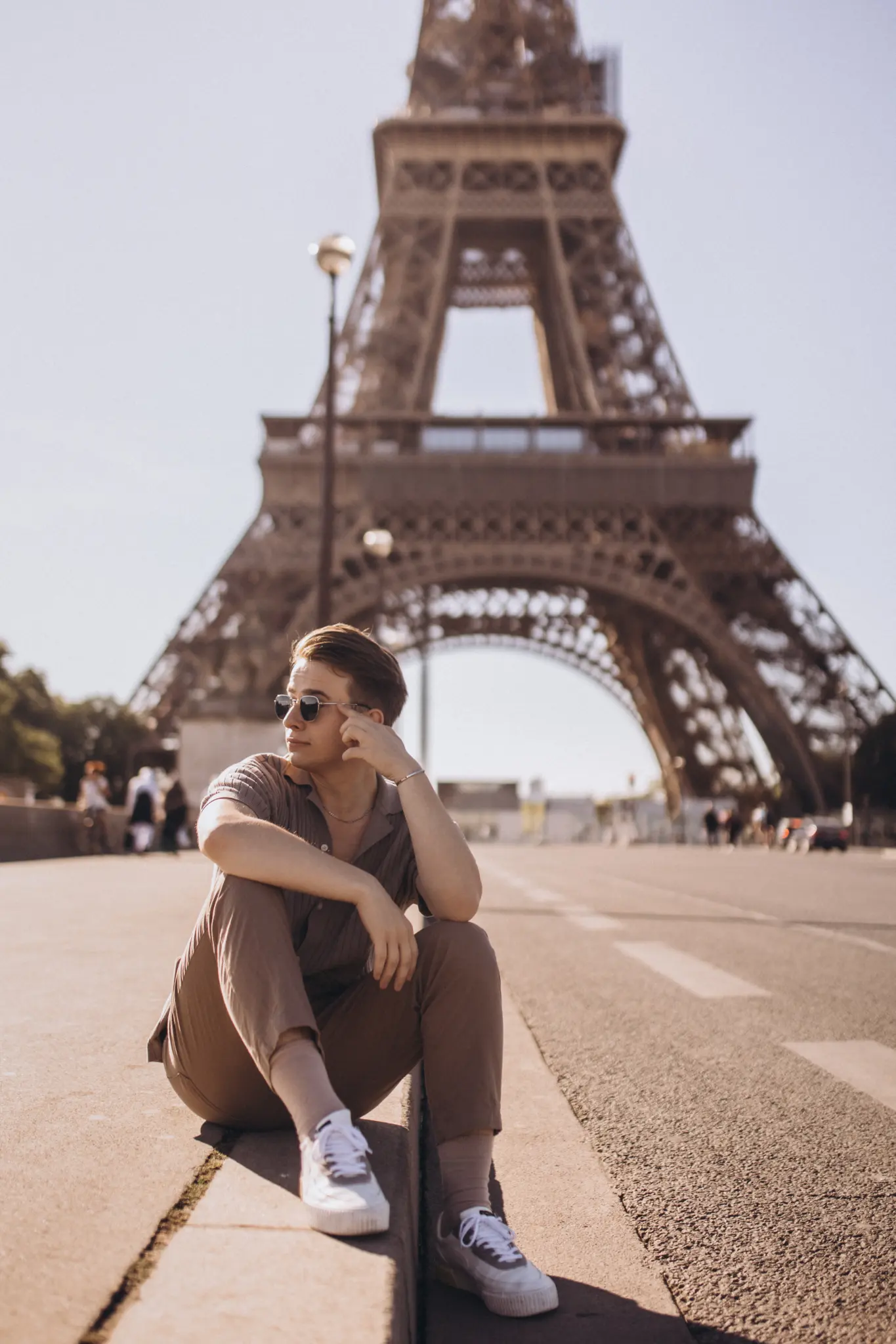 A young man seated on the esplanade at the foot of the Eiffel Tower in bright daylight, the landmark close behind.