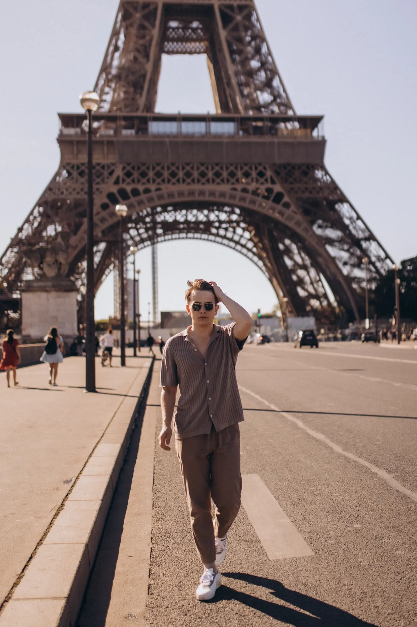 A young man walking across the open esplanade toward the camera, running a hand through his hair with the Eiffel Tower behind him.