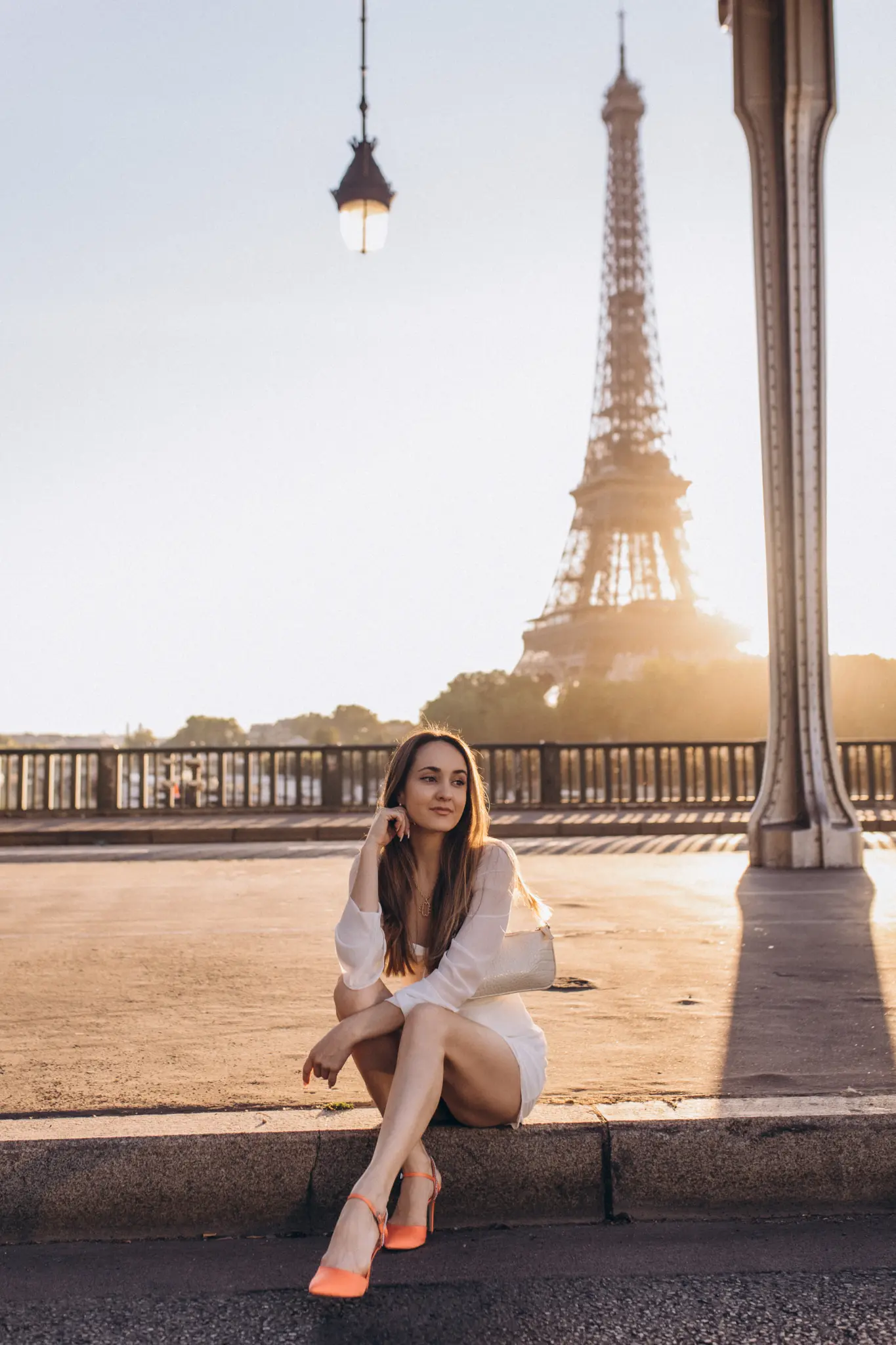 A woman in a white dress seated on the Bir-Hakeim bridge, a steel viaduct column and the sunlit Eiffel Tower behind her.