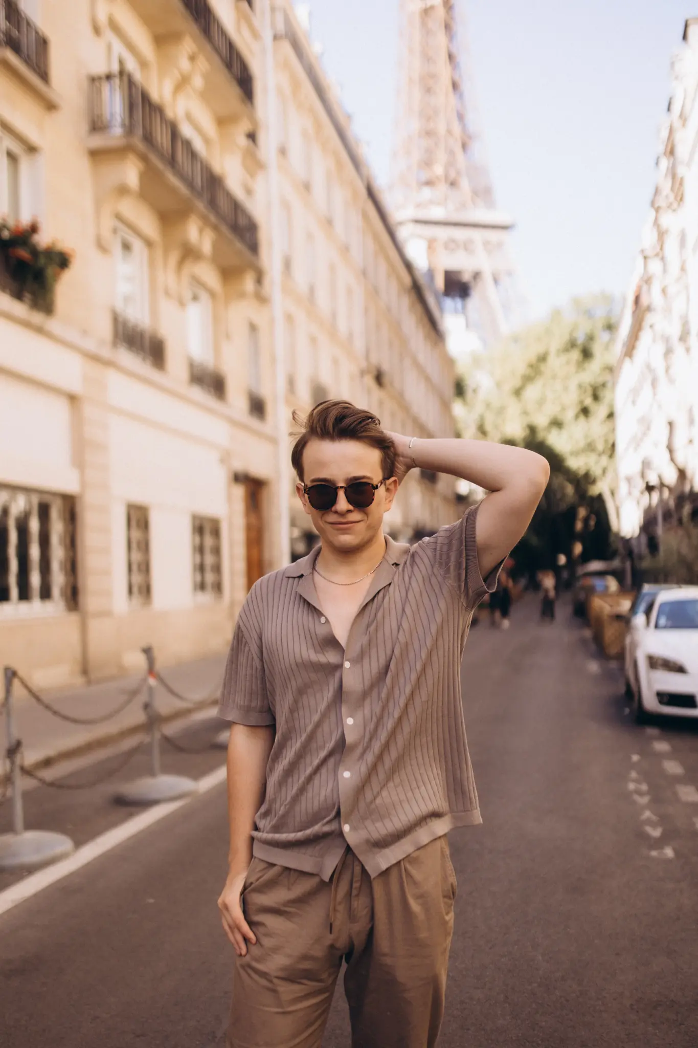 A relaxed male street-style portrait in Paris, a young man in sunglasses and a beige knit shirt with the Eiffel Tower in the background.