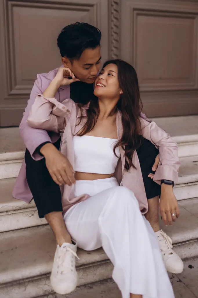A couple at their Paris engagement session sitting on stone steps and gazing at each other, the woman in white laughing softly, an engagement ring on her hand