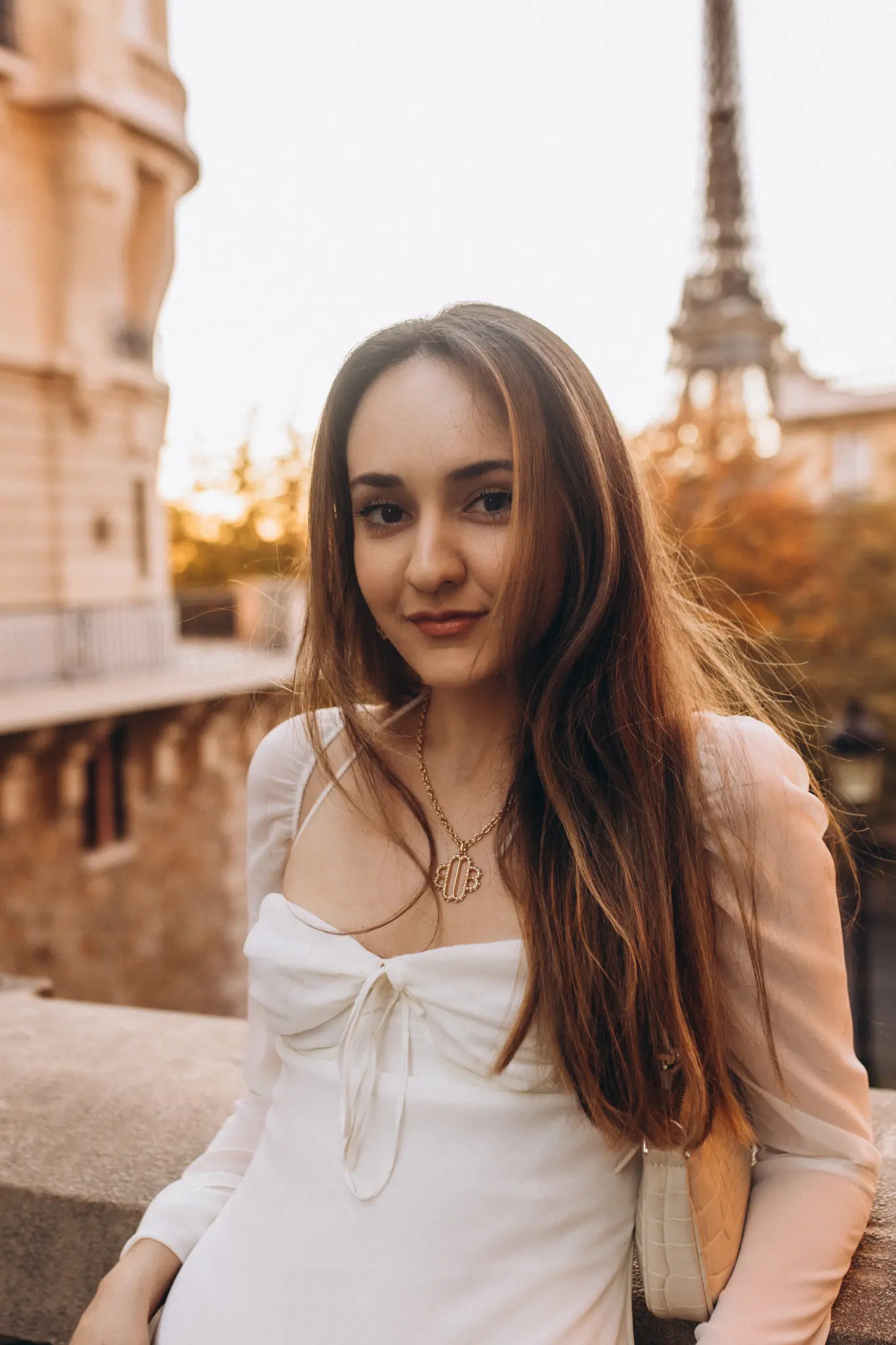 A woman in a white dress smiling gently at golden hour, the Eiffel Tower glowing in the warm light behind her.