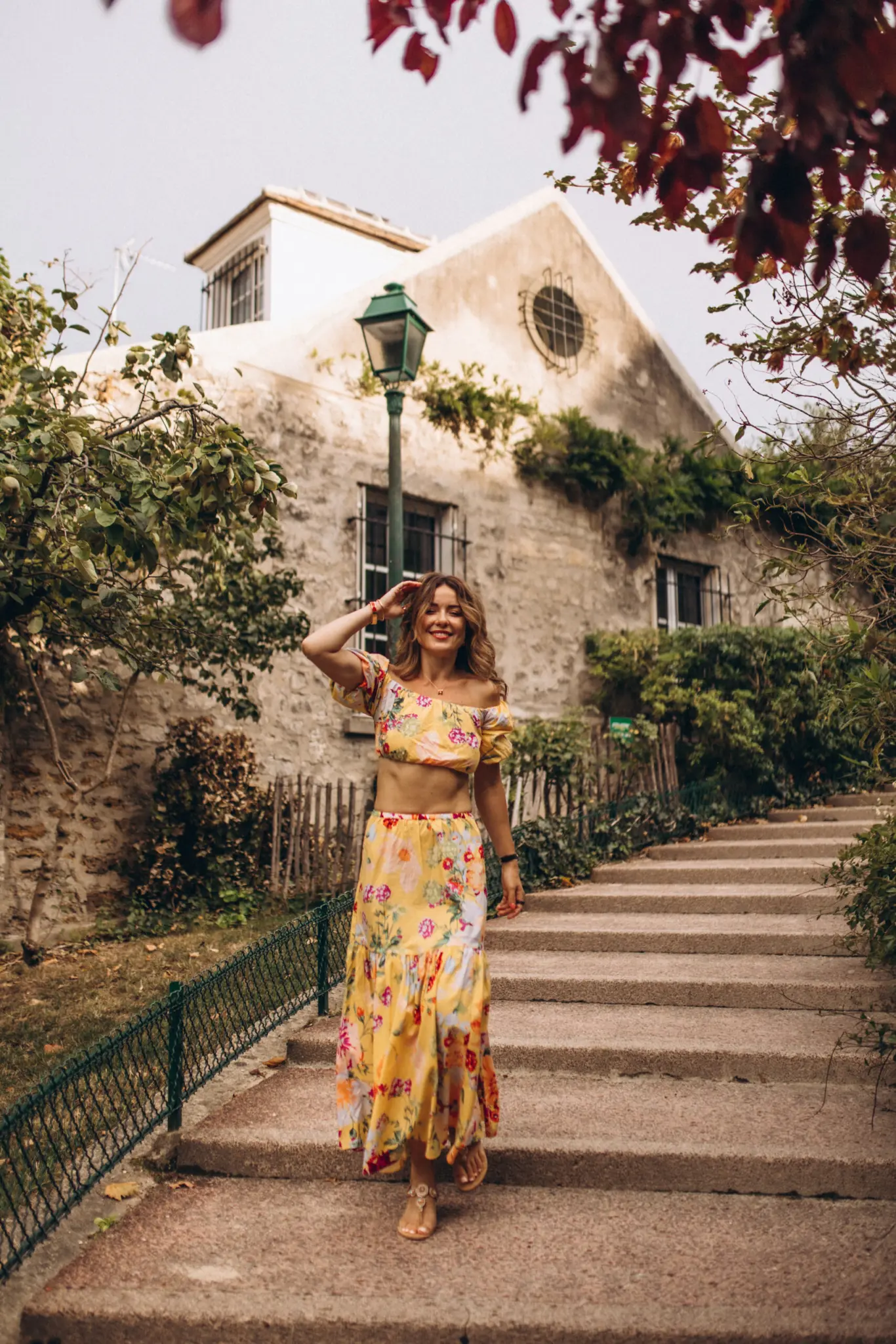 A photoshoot in Montmartre, a smiling woman in a yellow floral crop top and maxi skirt on old stone steps, one hand lifted to her hair