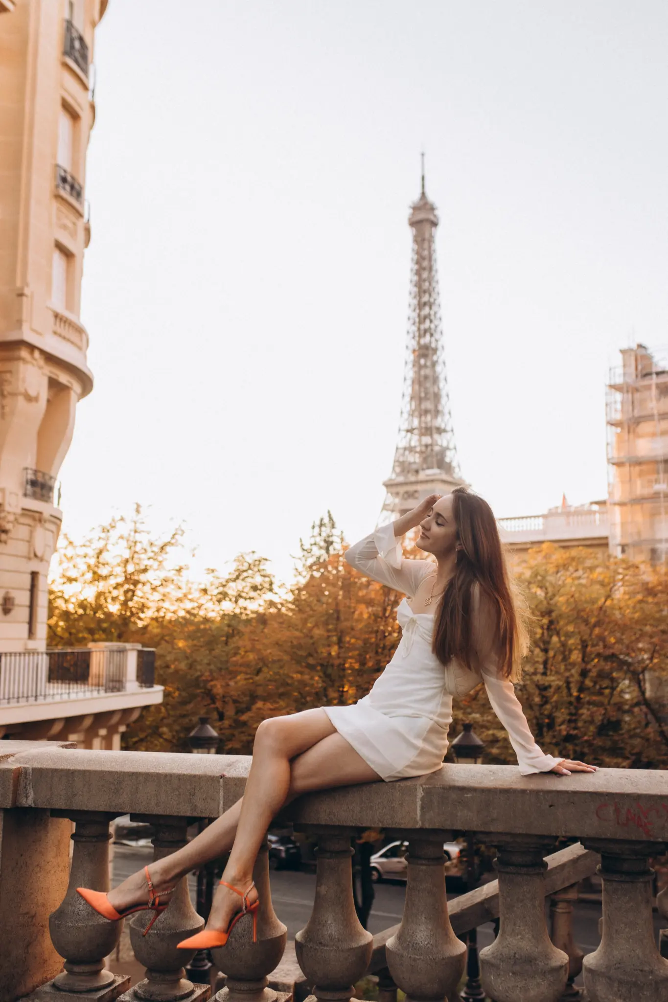 A woman in a white dress and orange heels sitting on a stone balustrade at golden hour, the Eiffel Tower rising behind her.