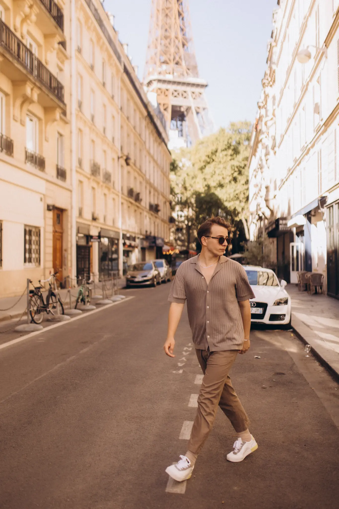 A young man in a beige shirt and chinos walking across a sunlit Parisian street, the Eiffel Tower rising ahead of him.