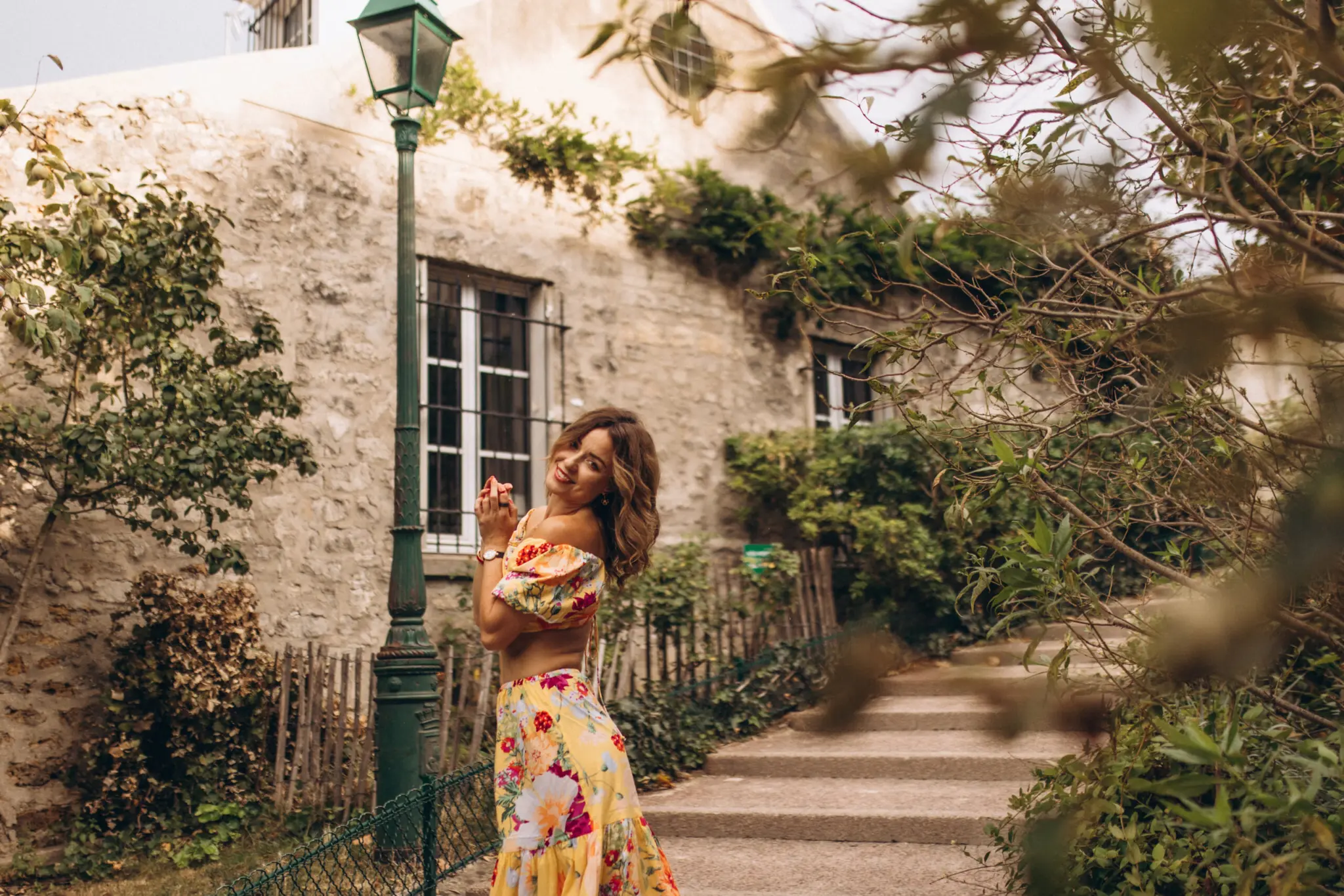Montmartre photoshoot in Paris, a woman in a yellow floral dress glancing back over her shoulder on a green, leafy Montmartre lane
