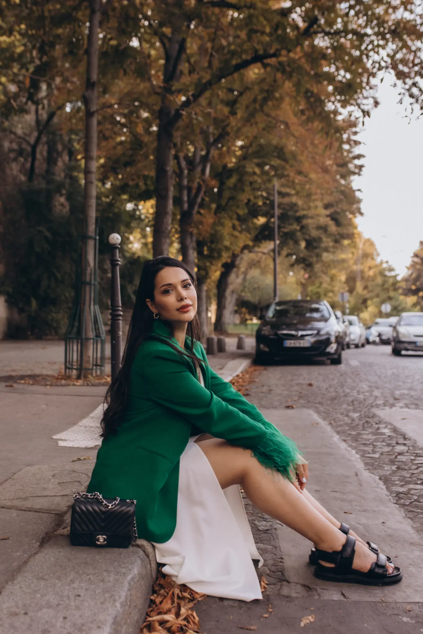 Paris portrait session, a woman in a green feathered blazer and white dress sitting on a curb of a tree-lined Parisian street in autumn