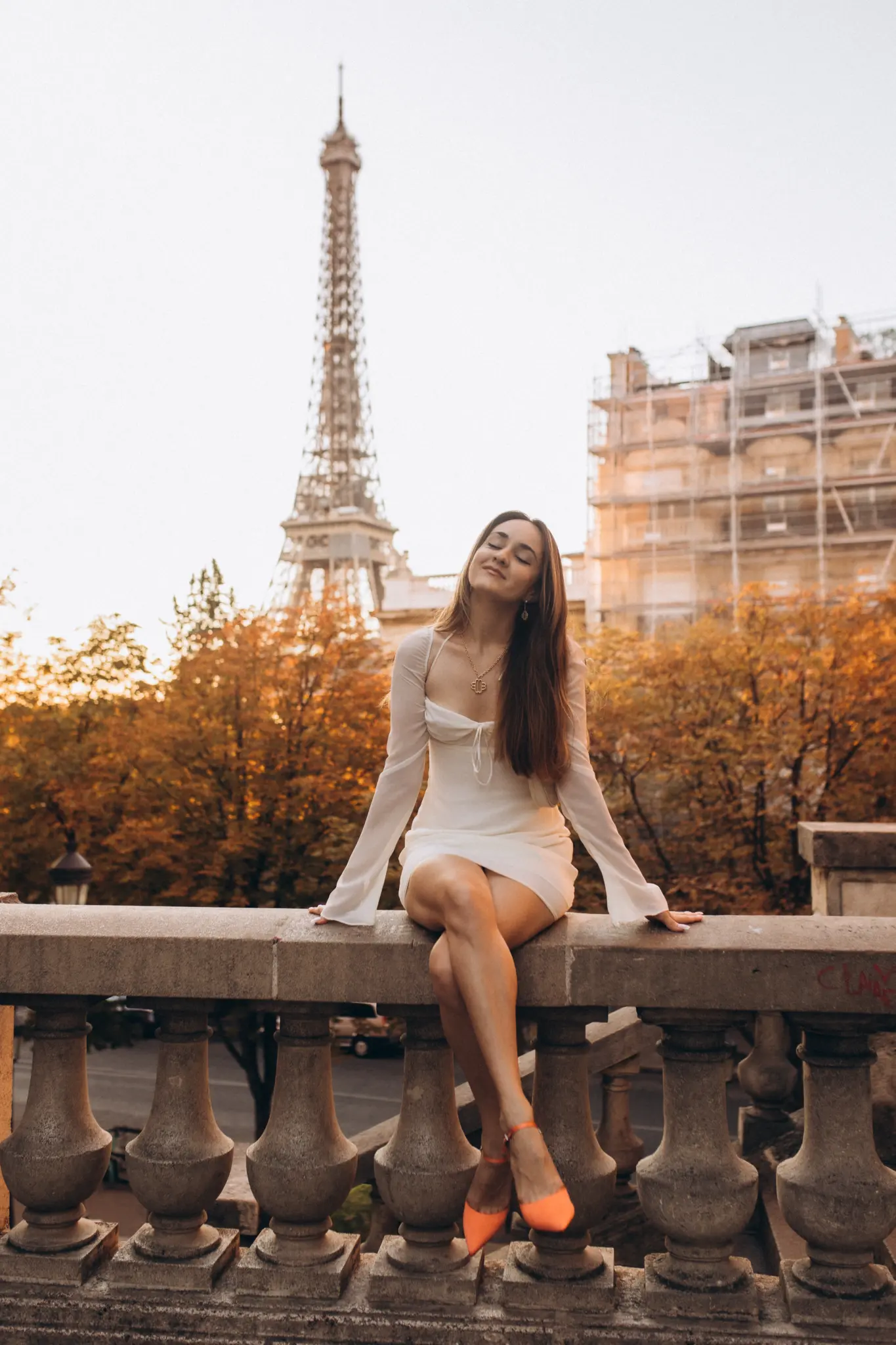 A woman in white seated on a balustrade among autumn foliage at golden hour, the Eiffel Tower behind her.