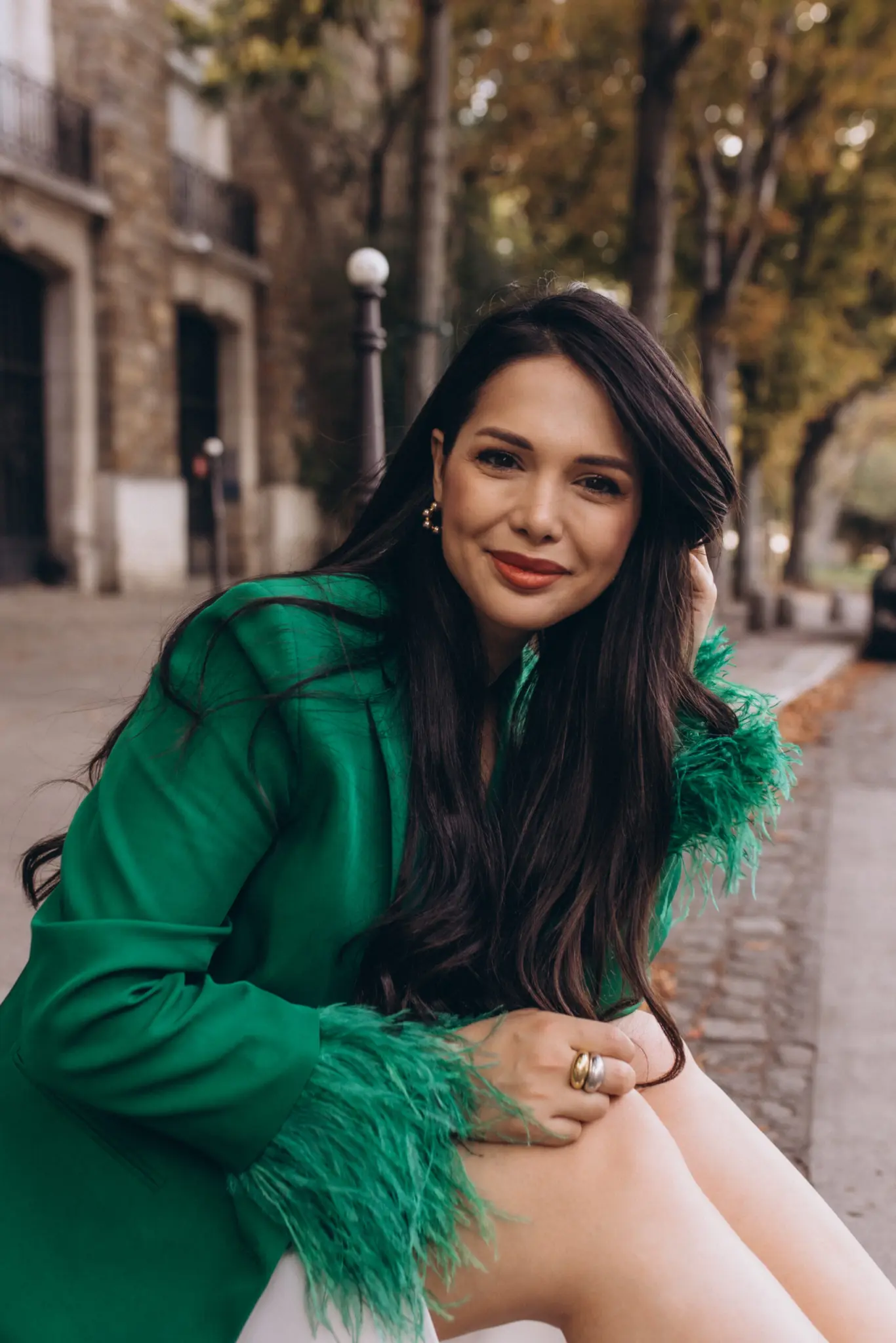 Paris portrait photoshoot close up, a smiling woman in an emerald feather-cuffed blazer with one hand lifted to her long dark hair