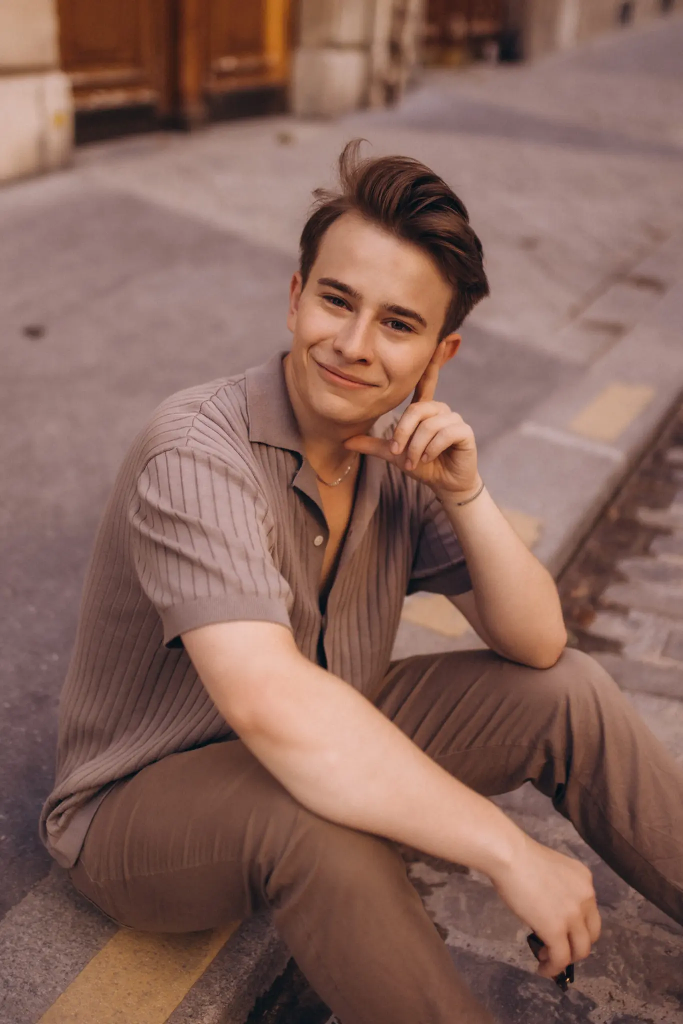 Paris street photography portrait, a young man sitting on a curb with his hand to his cheek, smiling warmly in soft light