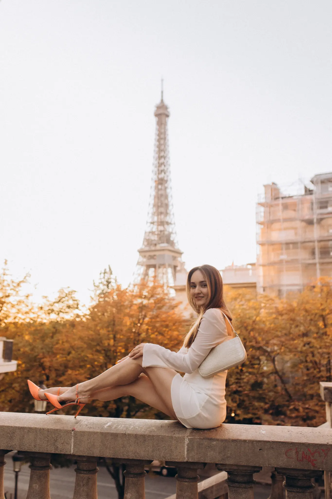 A woman in a white dress and orange heels sitting on a stone balustrade, autumn trees and the Eiffel Tower behind her.