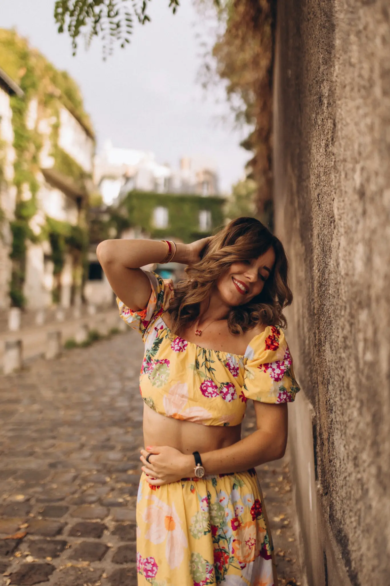 Summer Montmartre photoshoot in Paris, a woman in a yellow floral set smiling and running a hand through her hair against an old stone wall on a cobblestone lane