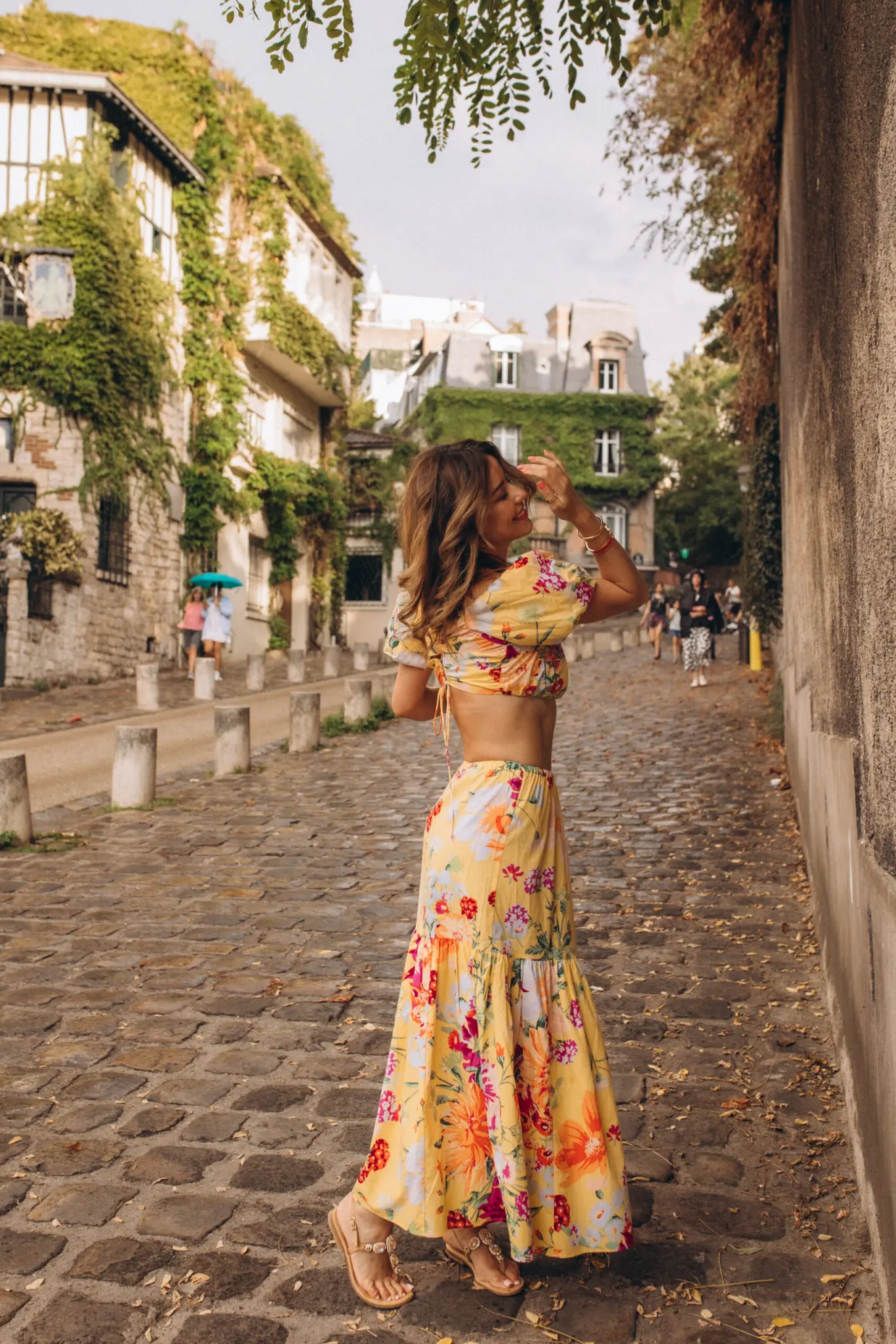 Montmartre photoshoot in Paris on Rue de l'Abreuvoir, a woman in a yellow floral two-piece walking away down the ivy-lined cobblestone lane, one hand lifted to her hair