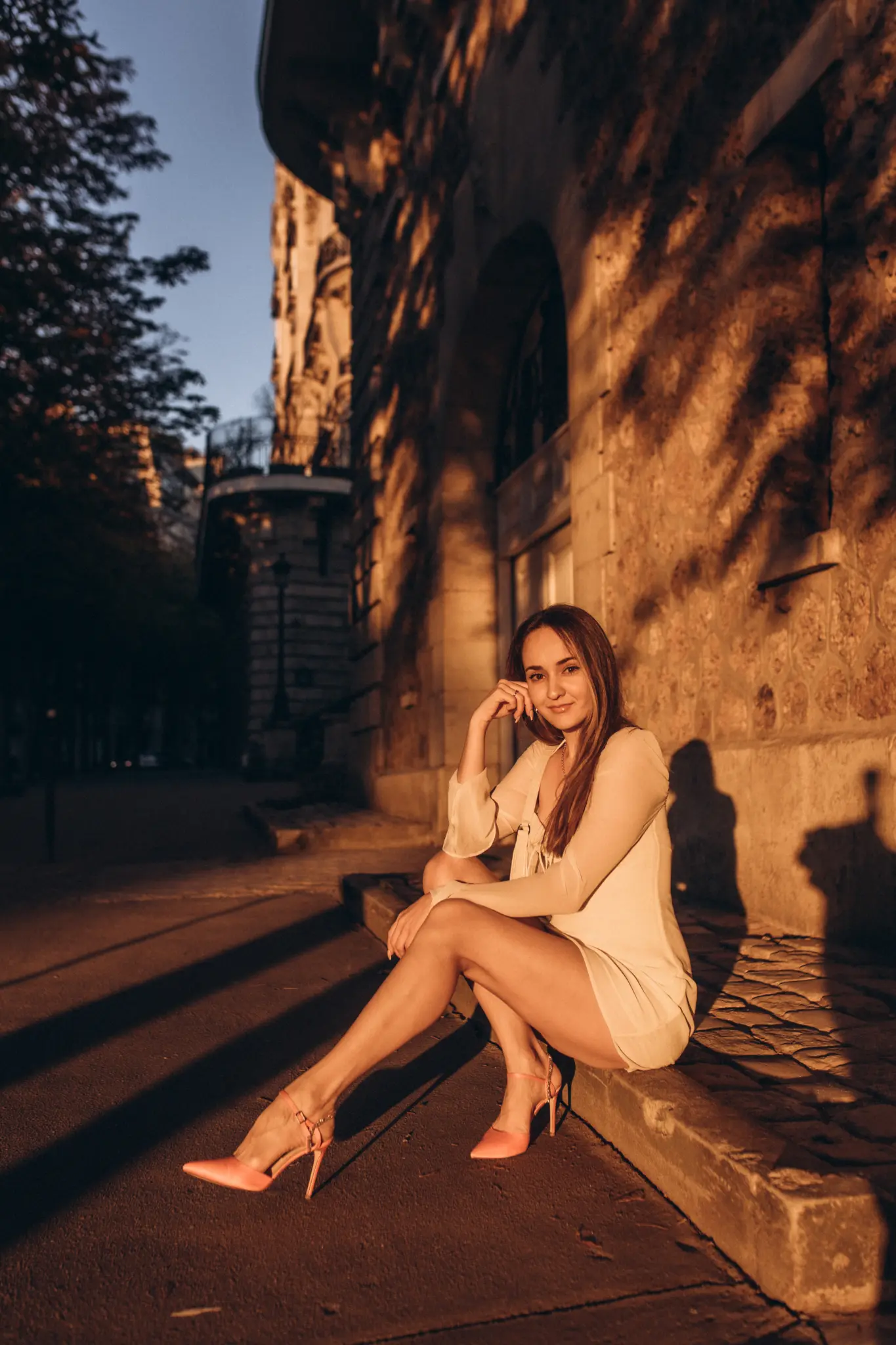 A woman in a white dress sitting on a stone step against a sunlit Parisian wall, warm light and soft shadows around her.