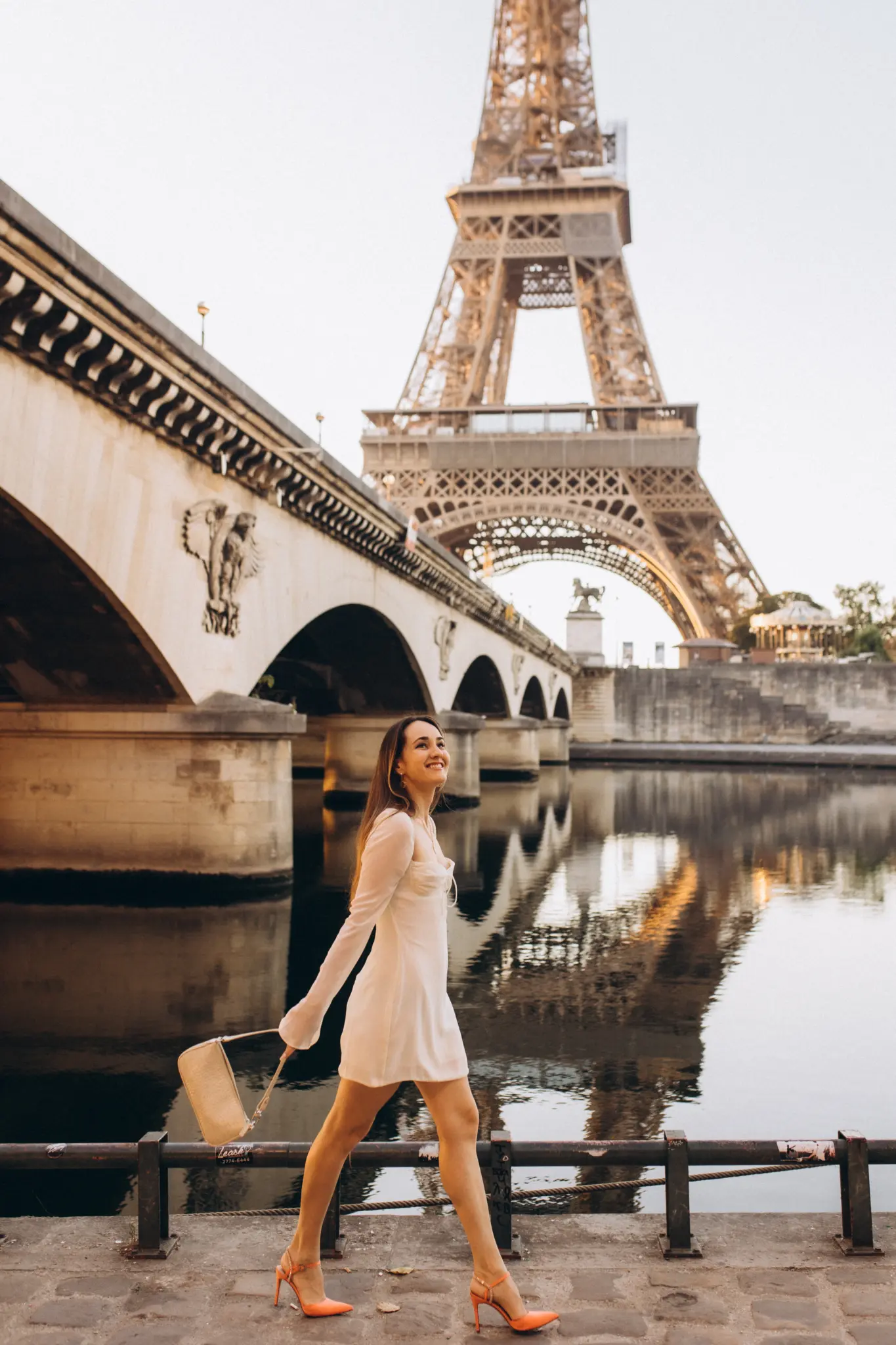 A woman walking along the Seine past a stone arch bridge, the Eiffel Tower reflected in the river.