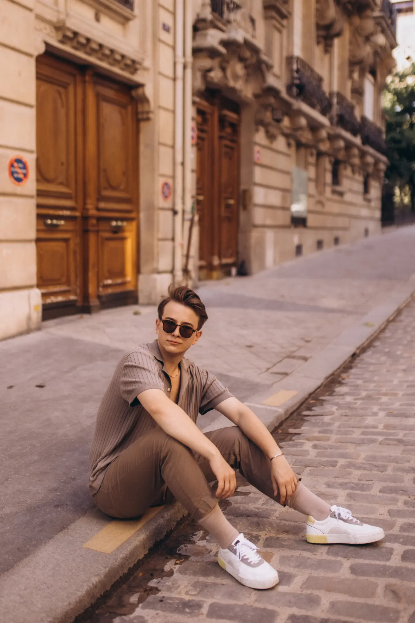 A young man in sunglasses sitting on a curb along a quiet cobblestone Paris street lined with wooden doors.