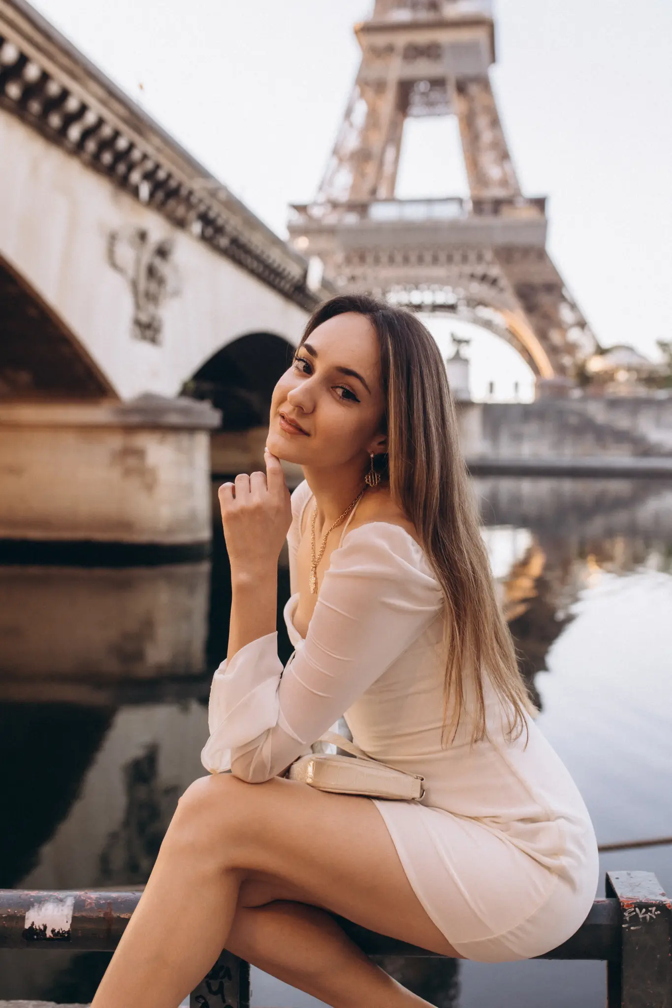 A woman seated by the Seine near a stone arch bridge with the Eiffel Tower rising close behind.