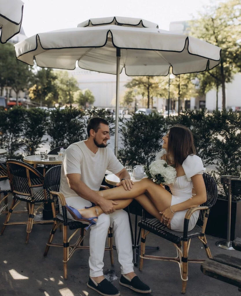 Newly engaged couple relaxing at a classic Paris café terrace under a white parasol during a Paris engagement photoshoot with Julia Litvin, the bride-to-be in white holding a bouquet of hydrangeas