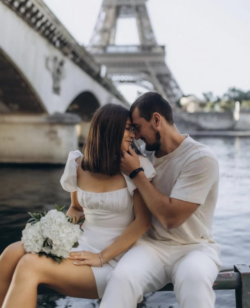 Tender newly engaged couple in white touching foreheads at their Paris engagement session, the bride-to-be holding a white hydrangea bouquet by the river - photo from Paris Engagement