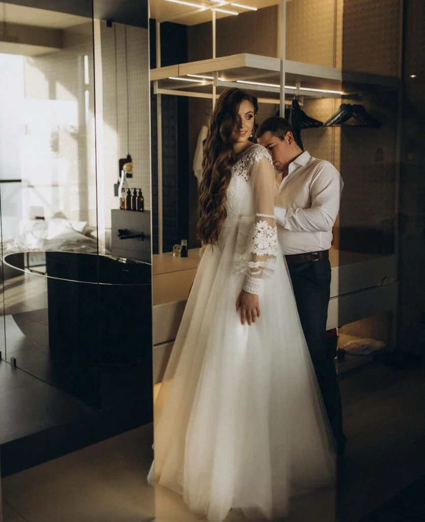 Newly engaged couple in an elegant hotel suite, the bride-to-be in a flowing white lace gown as her fiancé rests his head tenderly on her shoulder during a Paris engagement photoshoot