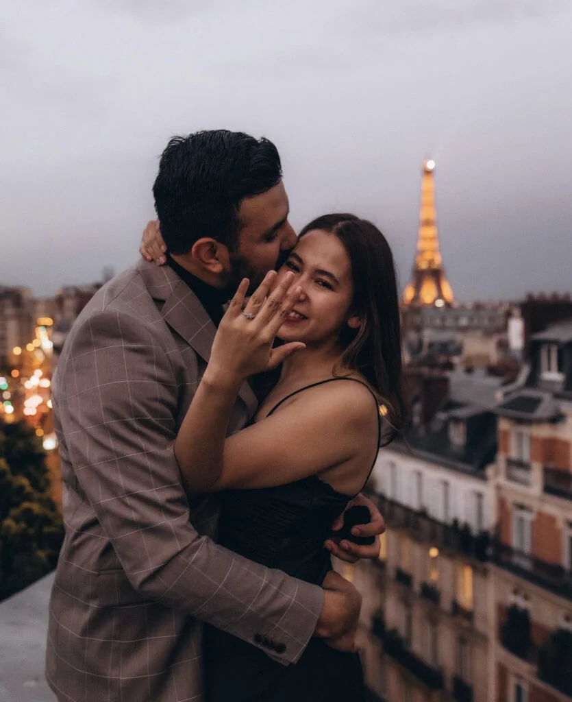 Woman shows off her engagement ring as her partner kisses her cheek, Eiffel Tower glowing behind