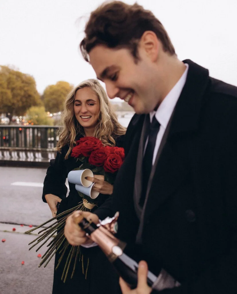 Newly engaged couple laugh together as the man opens champagne and the woman holds red roses