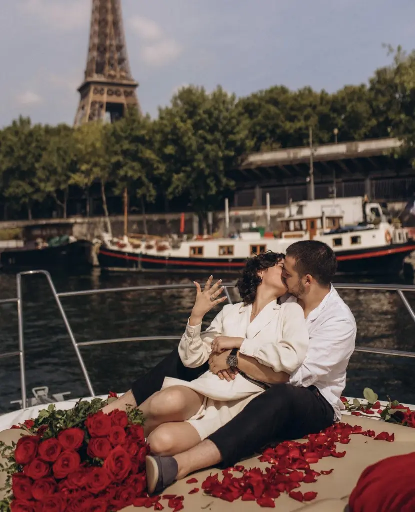 Couple kiss on a rose-covered boat as the woman shows her engagement ring, Eiffel Tower behind