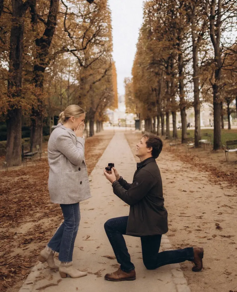 Man kneels with the ring on a leaf-covered path as his partner gasps, hands to her face