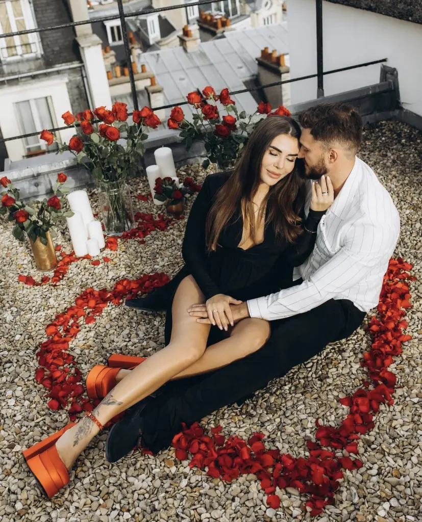 Newly engaged couple embrace on a rooftop among red roses, petals and candles, ring on her hand