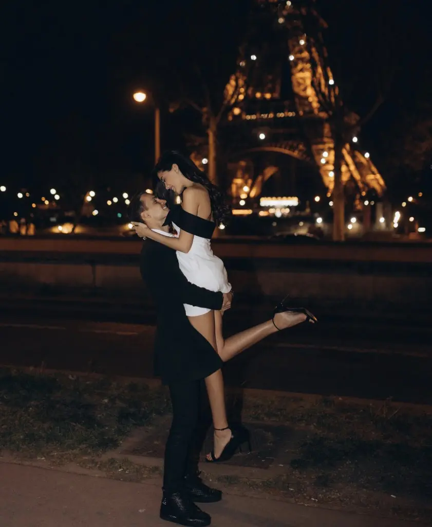 Man lifts his partner joyfully in his arms at night as the illuminated Eiffel Tower glows behind