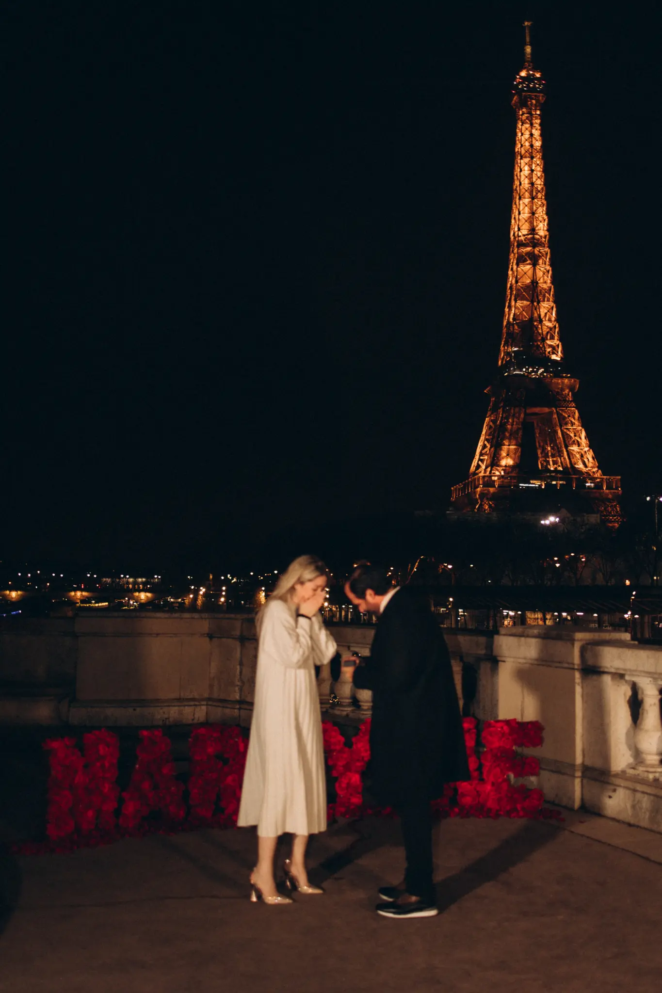 Woman covers her face in tears as her partner proposes beside rose letters, Eiffel Tower glowing