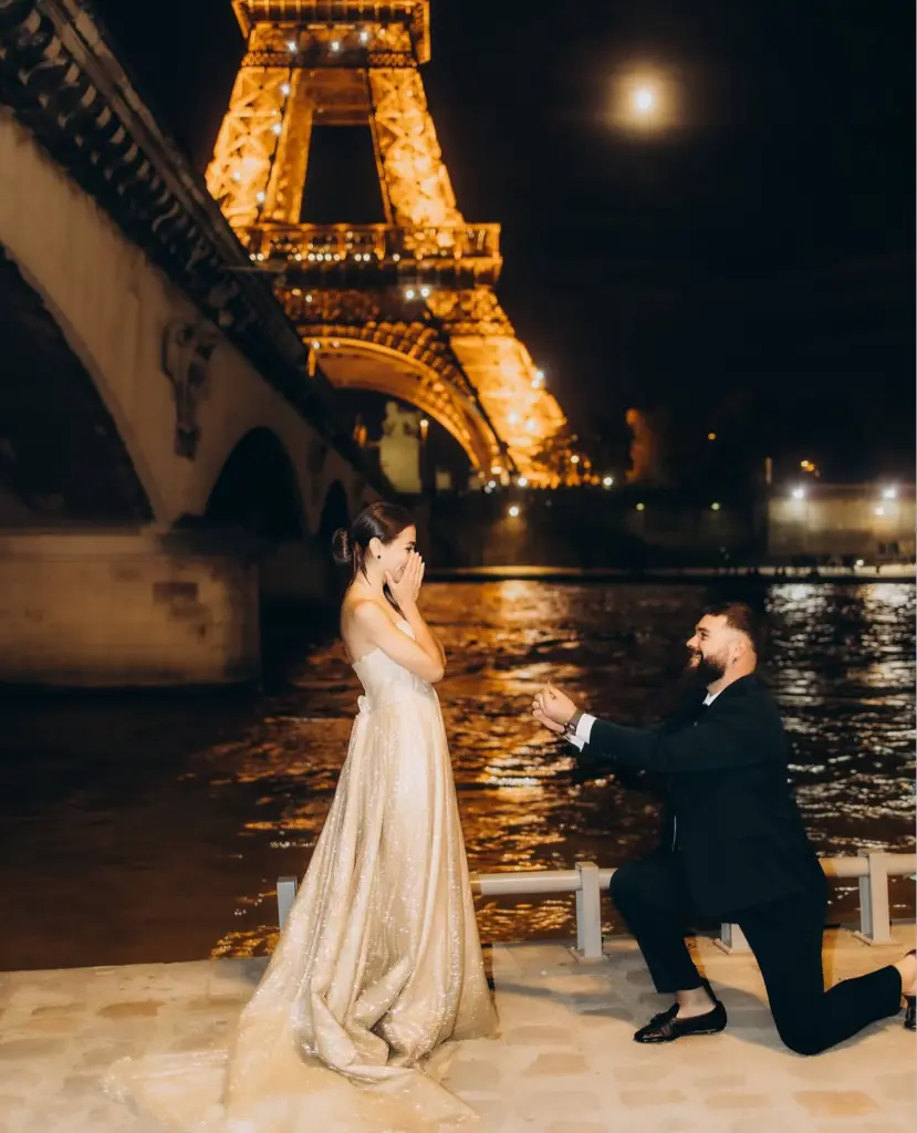 Her hands went to her face and stayed there. That is the frame, and it lasts about a second and a half: the river burning gold behind them, the moon hanging over the bridge like a lamp someone forgot to switch off, and one person completely undone by another. I photograph couples in Paris for exactly this second, and I have never once managed to breathe through it.