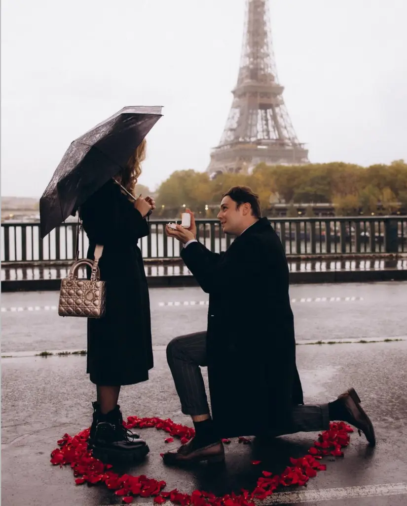 Couple photoshoot in the rain on a Seine bridge, a man kneeling with a ring box inside a heart of red rose petals, by Julia Litvin