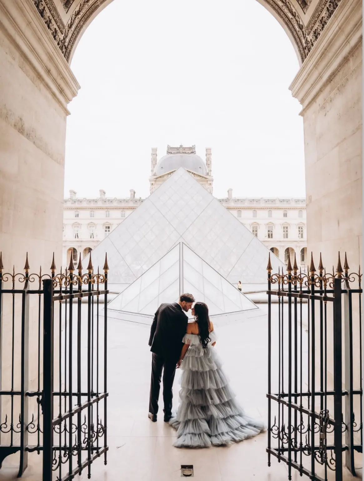 I waited for the courtyard to empty and shot from deep inside the passage, so the stone arch and the gilded gates would close around them like a proscenium. Two people, foreheads almost touching, a river of grey tulle pooling on the paving, and behind them the pyramid glowing white against a colourless sky. The Louvre holds centuries of paintings about love. That morning it held one more.
