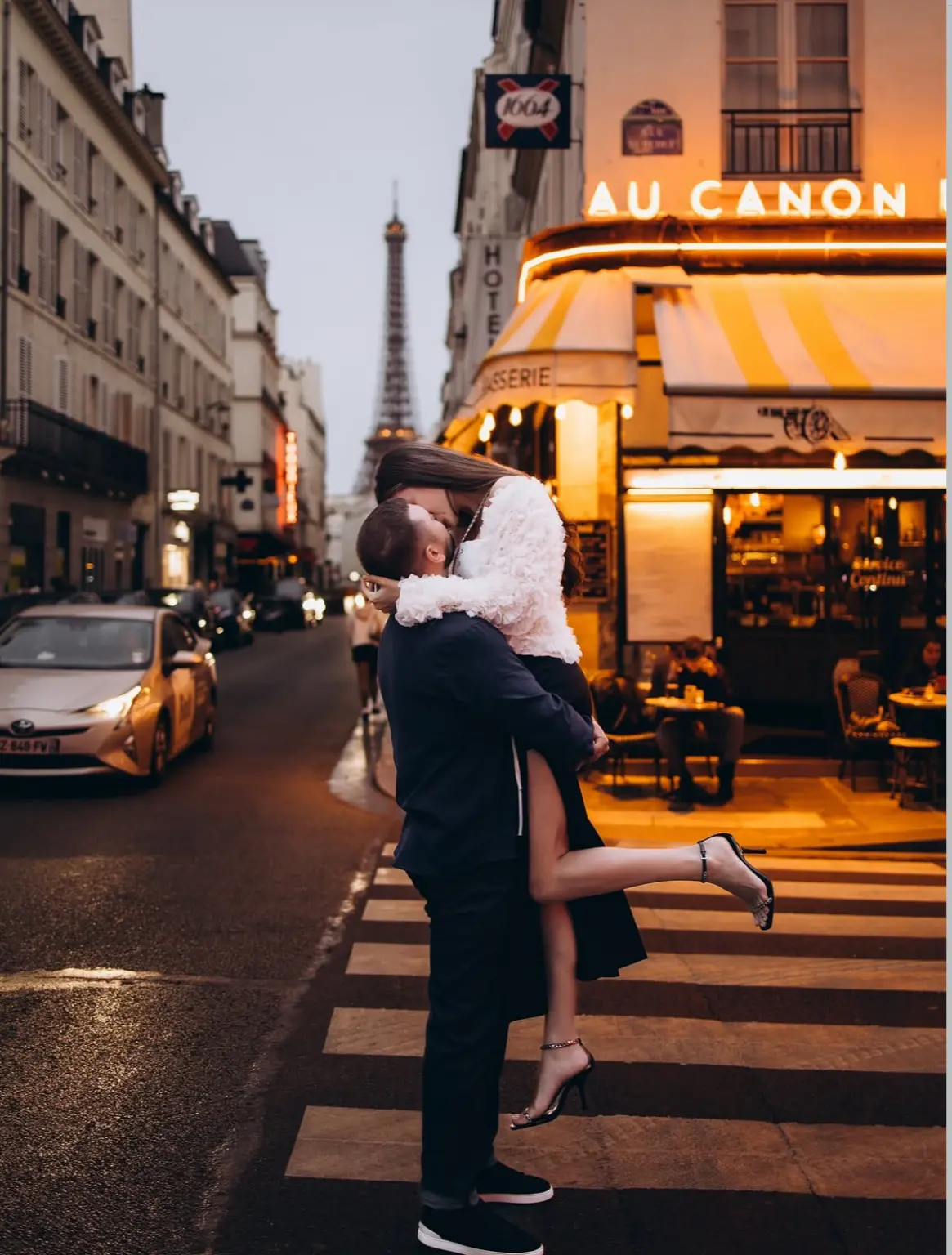 Paris couple photoshoot at dusk, a man lifting a woman for a kiss on a crosswalk beside a glowing corner brasserie, by Julia Litvin