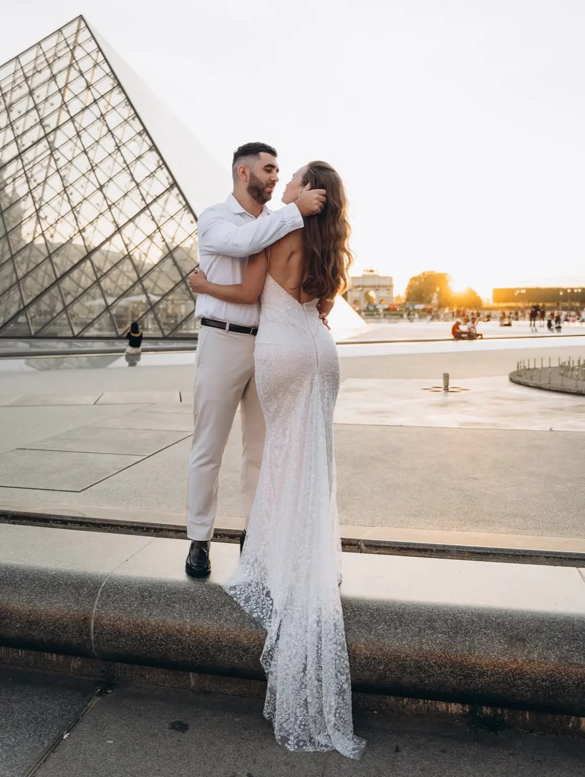 Couple photoshoot at the Louvre pyramid at sunrise, a woman in a lace gown and a man in white by the fountain rim, by Julia Litvin