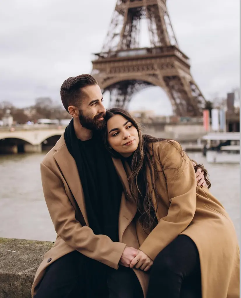 Professional couple photoshoot in Paris by the Seine, a couple in camel coats sitting on a stone parapet in winter, by Julia Litvin