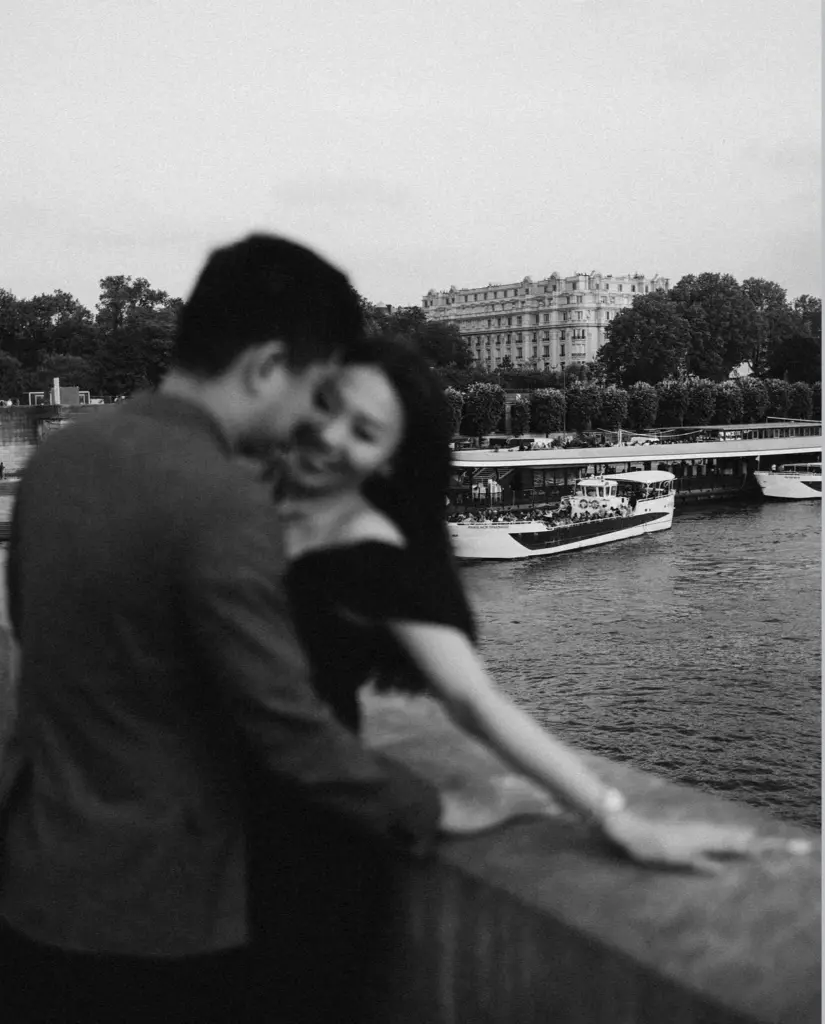Black and white Paris couple photoshoot on a bridge over the Seine, a couple embracing on the parapet as a boat passes, by Julia Litvin