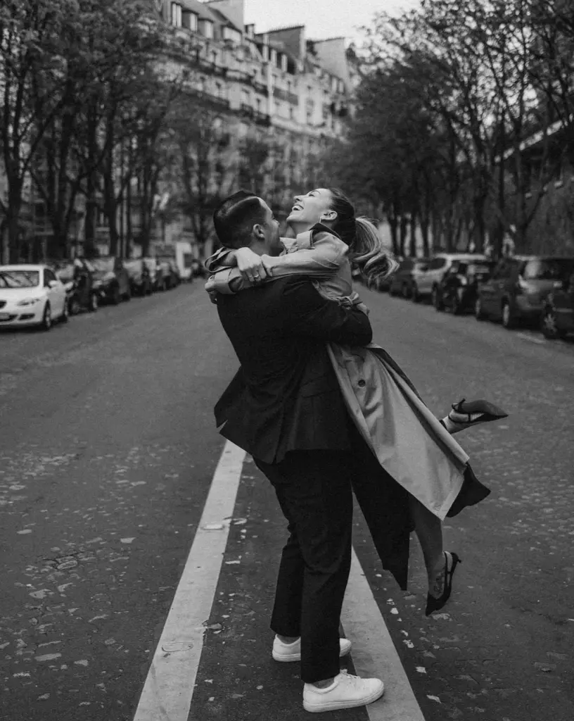 I stood right on the white line with the boulevard running away behind them, the bare plane trees leaning in from both sides, and told them one thing: pick her up. Her head went back, her heels flew, the trench opened like a sail, and the laugh that came out of her was not for me and not for the camera. Twelve seconds later a car came. Worth it.