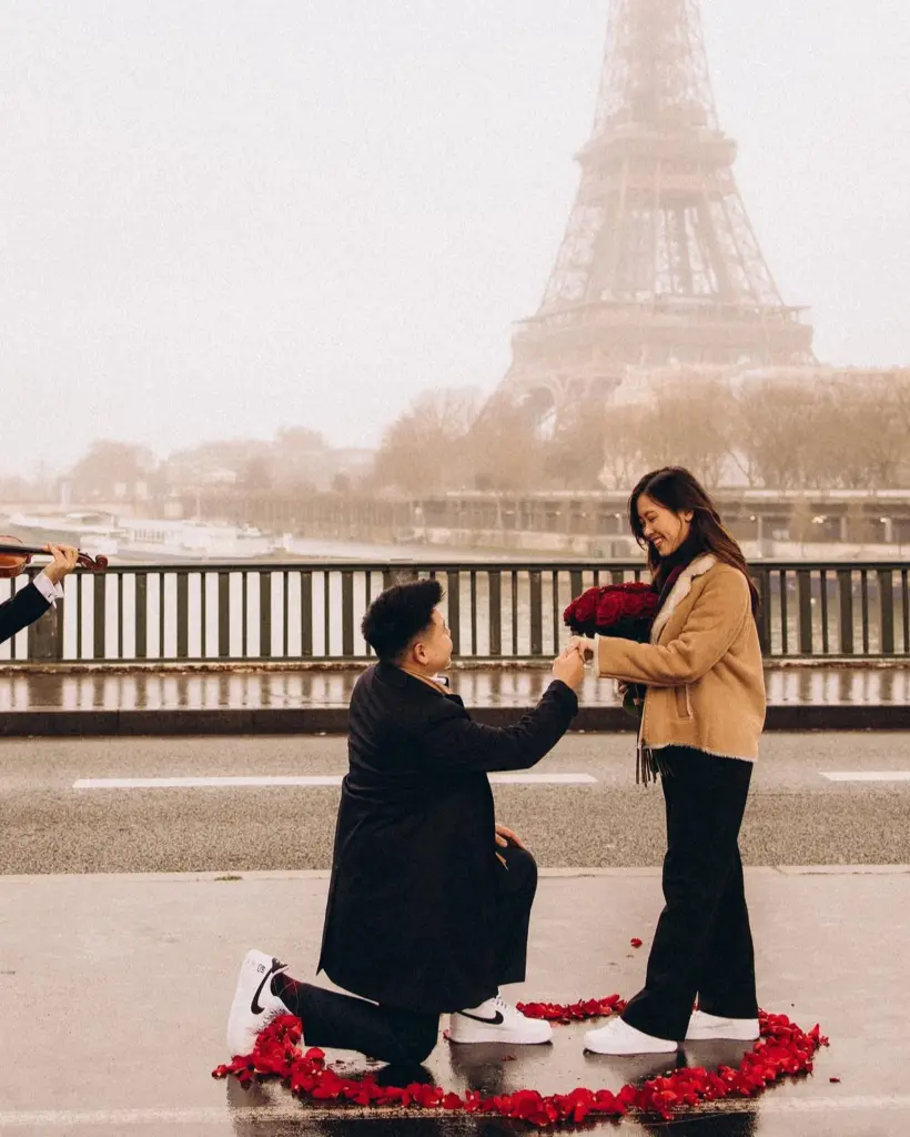 Couple on a Paris bridge in winter fog, a man kneeling with red roses inside a heart of petals, by couples photographer Julia Litvin