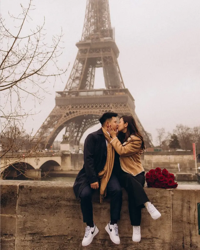 Couple kissing near Eiffel Tower with flowers - Paris couple photoshoot in winter fog, a couple kissing on a stone parapet above the Seine with red roses beside them, by Julia Litvin