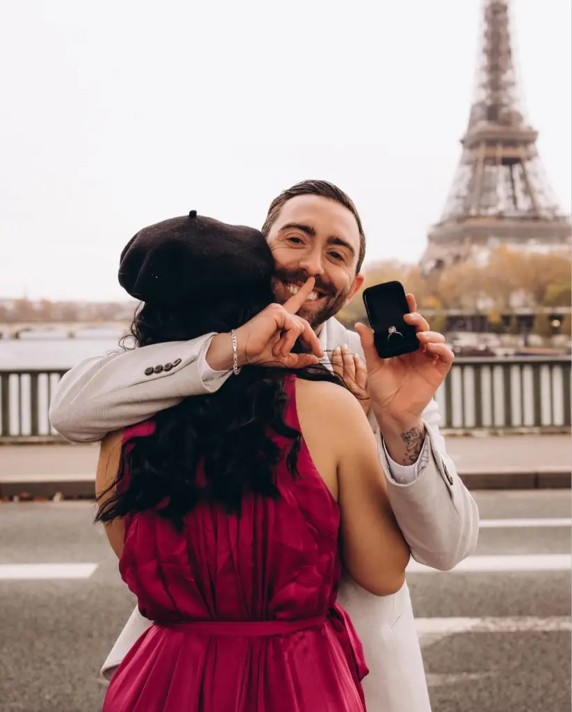 Couple photoshoot in Paris, a man embracing a woman in a raspberry dress while holding up a ring box behind her, by Julia Litvin