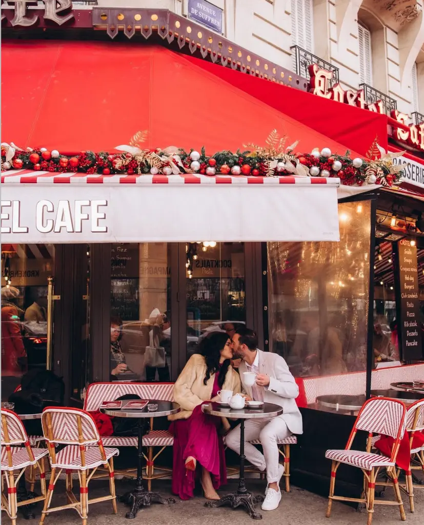Couple photoshoot on a Paris café terrace, a couple kissing at a small table under a red awning, by Julia Litvin