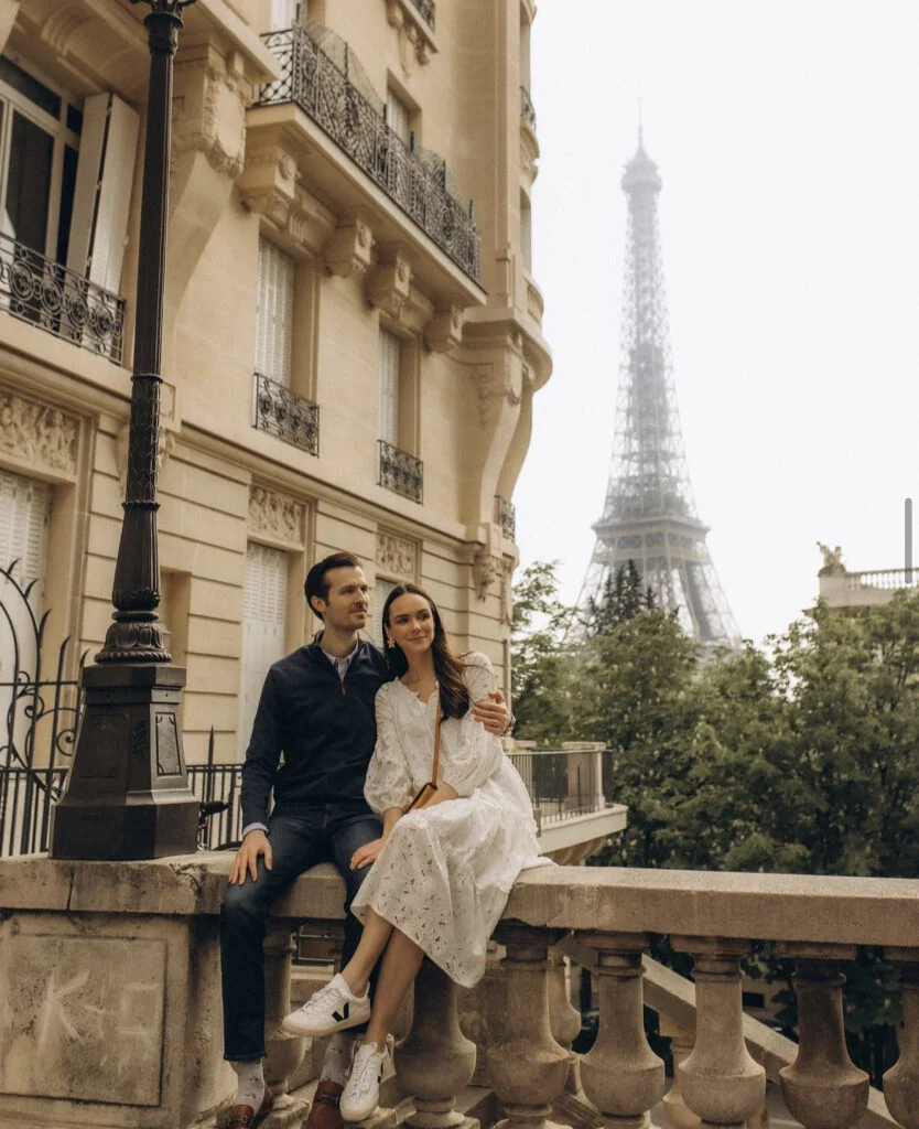 A couple seated on a stone balustrade on Avenue de Camoens with the misty tower behind them during an Eiffel Tower photoshoot